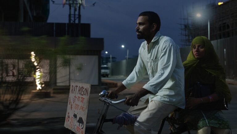 Man cycles past a sign labeled RAT POISON with a woman in a green headscarf riding on the back during the night.