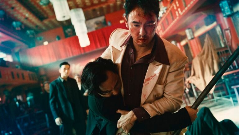 A bloodied man in a white suit grapples with an opponent inside a red-lit, multi-level theater.