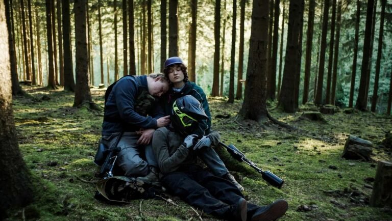 Three young people in paintball gear rest together on the mossy ground of a sunlit pine forest.