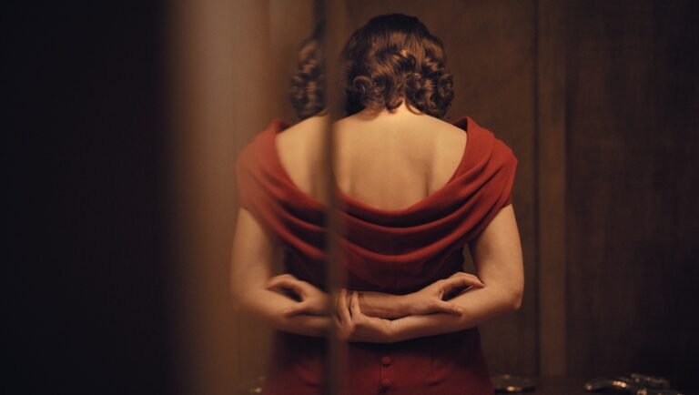 Actress Mathilde Arcel stands with her back to the camera, reflected in a mirror while wearing a red dress.