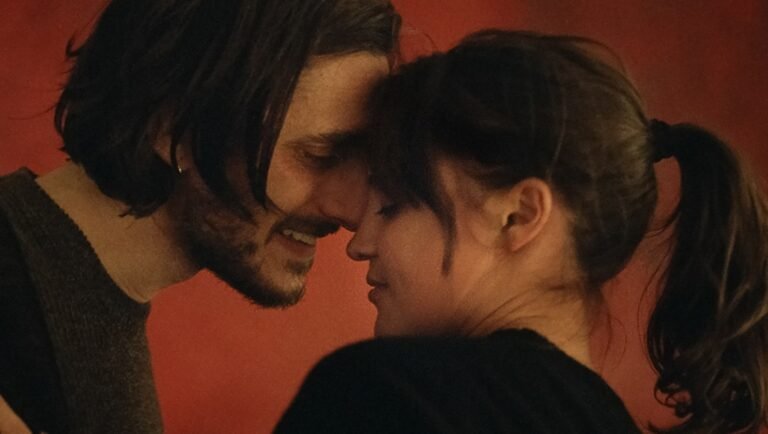 Luca Marinelli and Alicia Vikander touch foreheads affectionately in front of a warm red wall.