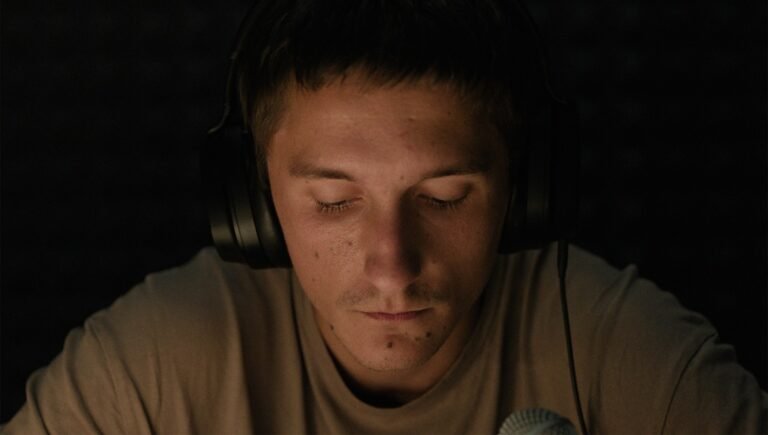 Arthur Aliiev sits with eyes closed while wearing large black headphones in a dimly lit, dark space.