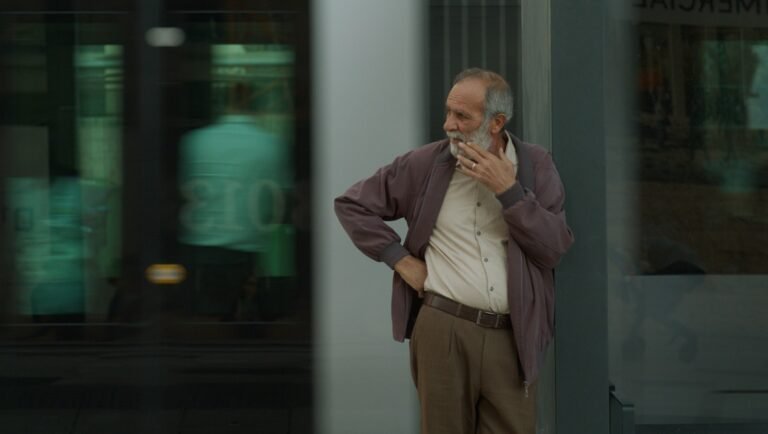 Middle-aged man with a gray beard wearing a maroon jacket and brown trousers touching his chin while standing outdoors.