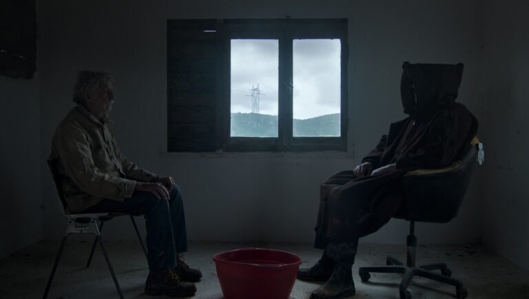 An older man sits facing a hooded figure in a dark room with a window overlooking a landscape and a red basin between them.