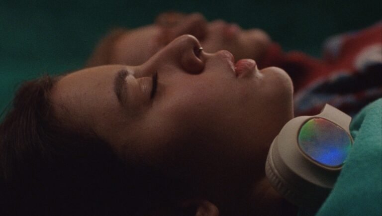 Close-up of a person with eyes closed wearing iridescent white headphones while lying on a dark green surface.