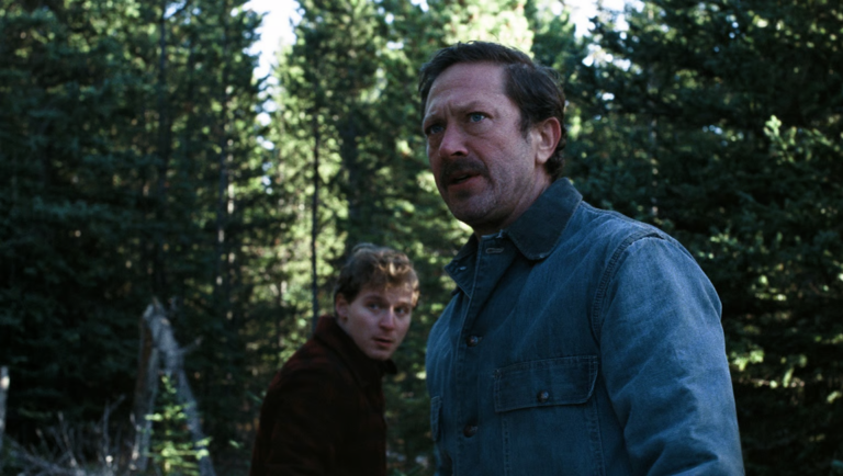 Two men standing in a dense forest, one in a blue shirt looking intently ahead, the other in a brown jacket behind him.