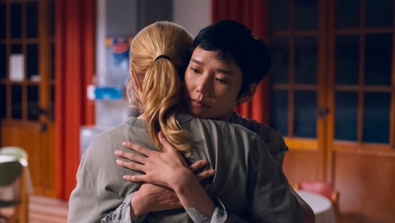 Two women embracing indoors, one with short dark hair resting her head on the other's shoulder, red curtains behind.