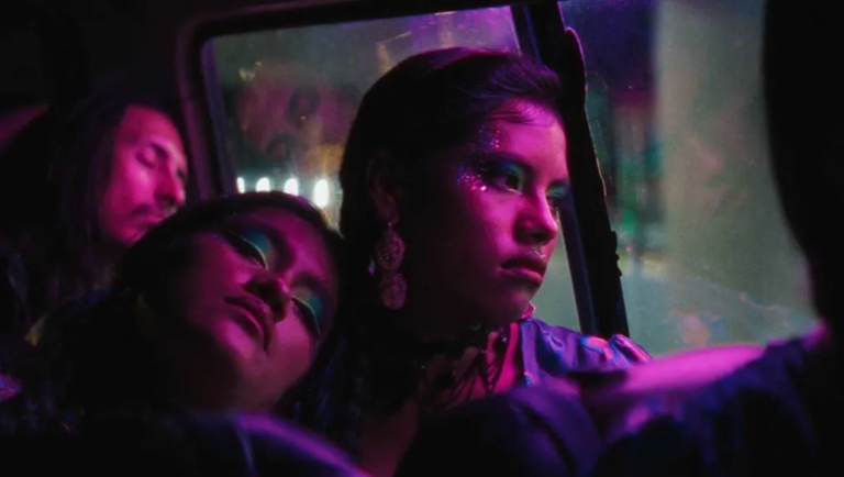 Three young women with glittery makeup sit inside a vehicle bathed in pink and purple neon light, one gazing out the window.
