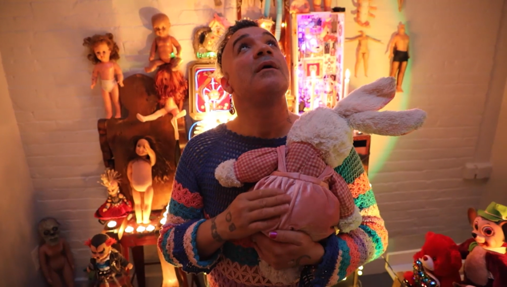 Person in a striped sweater holds a stuffed rabbit in a room filled with vintage dolls.