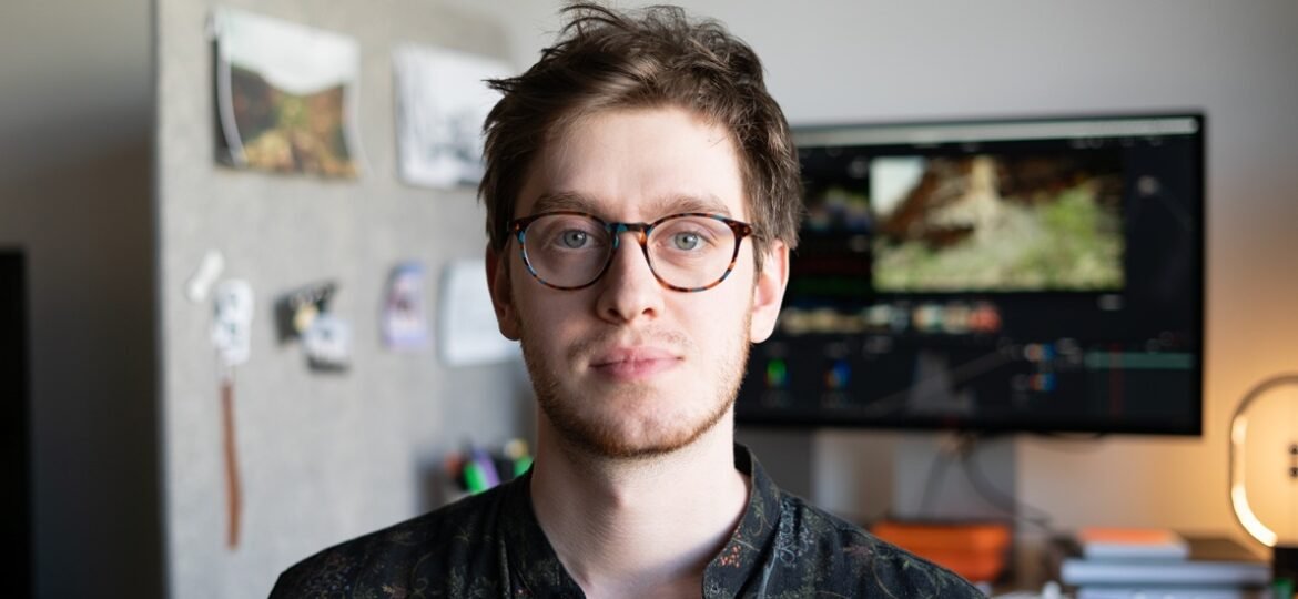 Bearded man in glasses and dark patterned shirt facing camera in a home office with a monitor behind him