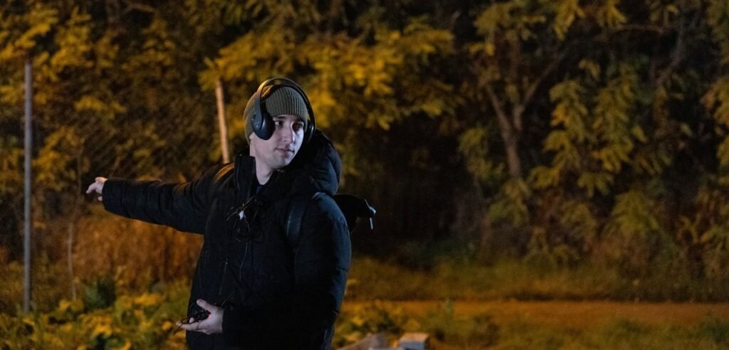 Man in black jacket and headphones pointing on set at night with trees behind him
