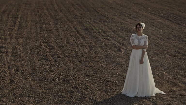 Bride in a white lace gown and veil standing alone in a vast plowed field at dusk.