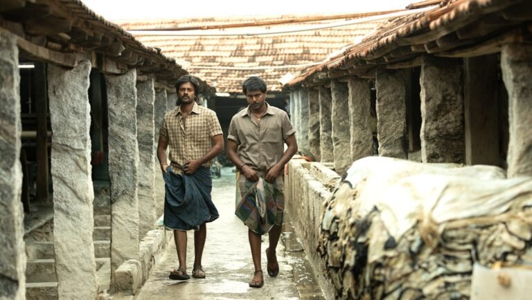 Two men in shirts and traditional lungis walk through a narrow, stone-pillared corridor with stacked hides.
