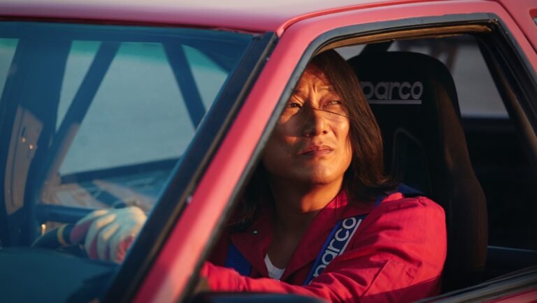 Long-haired man in a red racing suit sitting in a pink race car with a Sparco seat