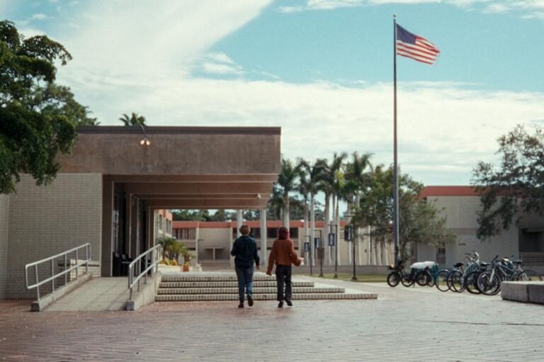 Two people walk up concrete steps toward a brick building under a large awning, with an American flag flying to the right.
