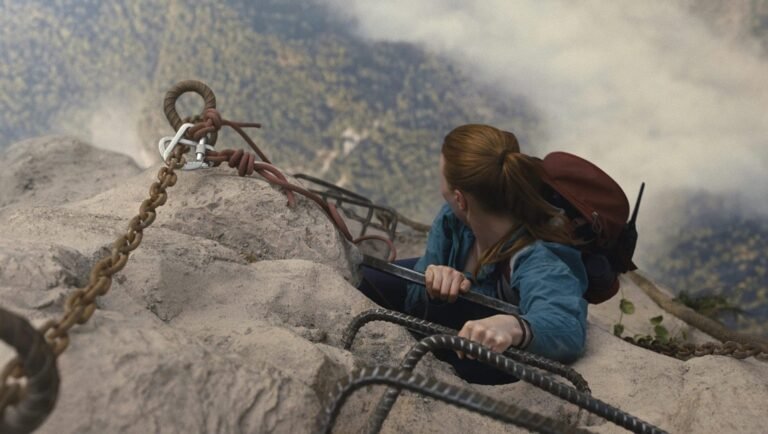 Woman with a ponytail and backpack climbs a steep rock face using metal rungs and a safety chain.