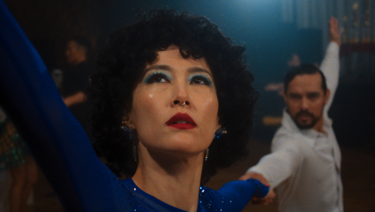Rinko Kikuchi in blue sequins with curly black hair and red lips dances beside a man in a white shirt.