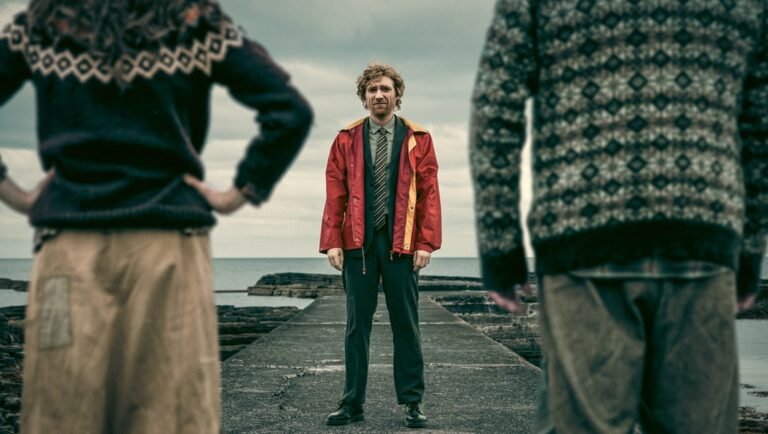 Man in red jacket and tie stands on a concrete pier facing two figures in patterned sweaters.