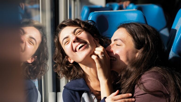 Two women laughing together while sitting on a train with blue seats in bright sunlight.