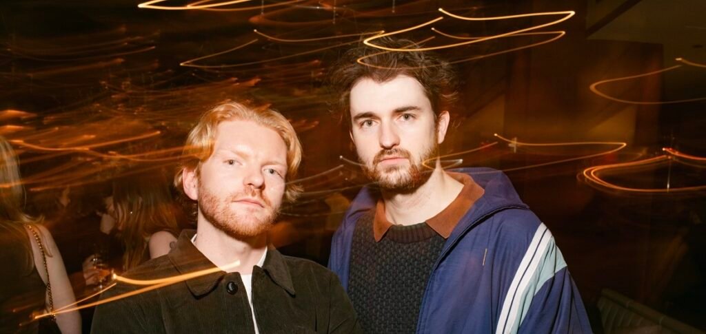 Two men with beards posing indoors at night with long-exposure light trails streaking across the background.