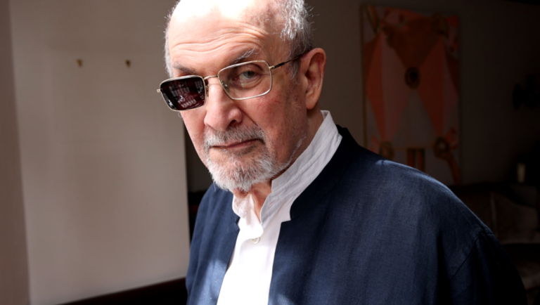 Salman Rushdie wearing glasses and a dark blue shirt looks directly at the camera with a serious expression.