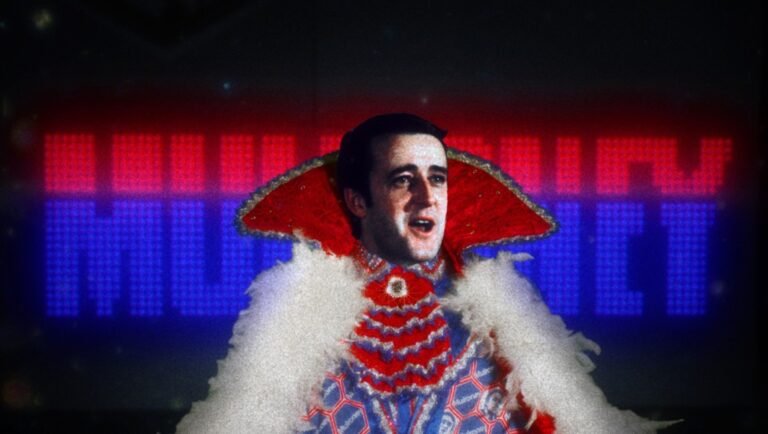 Man in an ornate red and white Elizabethan collar sings before a glowing LED screen reading MULRONEY.