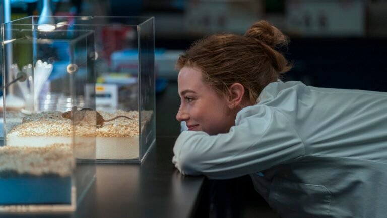 Smiling researcher in a lab coat leans on a bench to watch a mouse in a clear cage