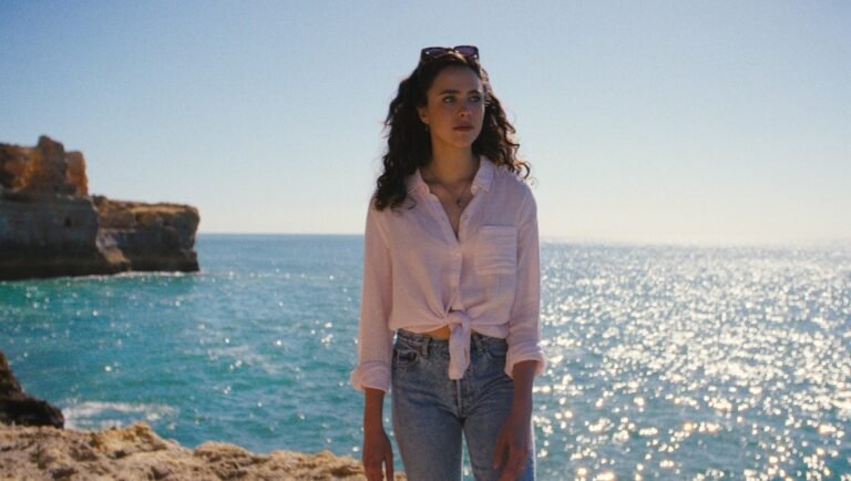 Woman with long dark hair stands on a rocky cliff, wearing a white knotted shirt and jeans, with sparkling ocean water and a rock formation behind her.