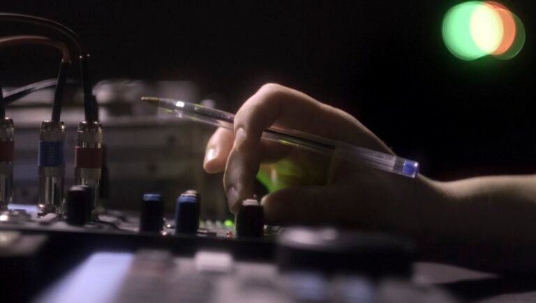 Hand holding a pen while adjusting a knob on a dark audio mixing console with glowing indicator lights.