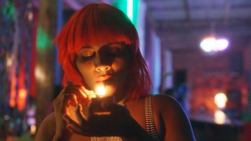 Woman with bright orange hair shields a glowing flame with her hand in dim neon light.