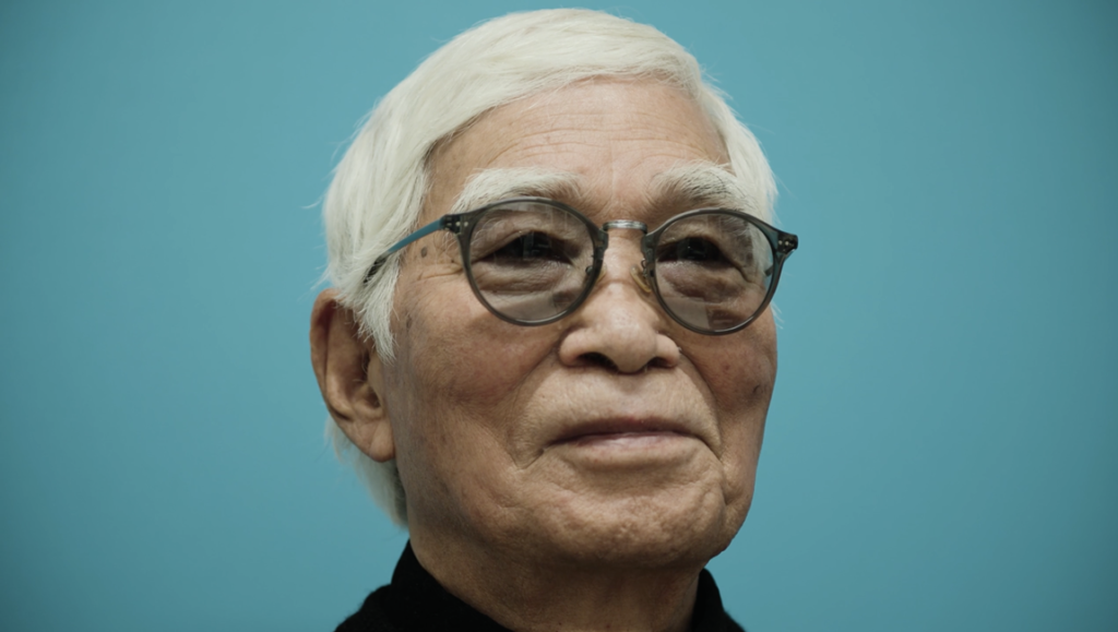 Elderly man with white hair and round glasses against a solid teal background.