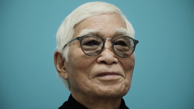 Elderly man with white hair and round glasses against a solid teal background.
