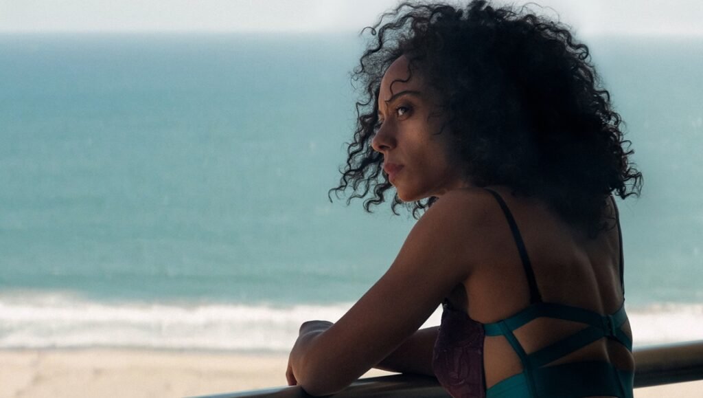 Woman with dark curly hair in a teal swimsuit looking back over her shoulder toward the ocean