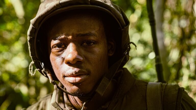 Close-up of a sweat-beaded soldier in a canvas helmet gazing intently through dense jungle foliage.