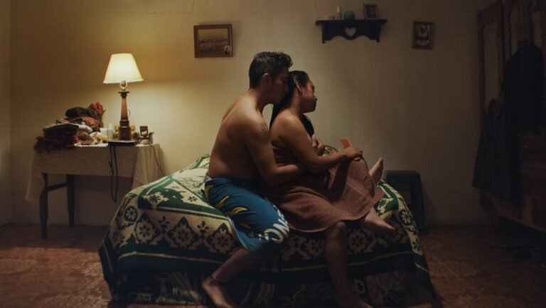 A man and woman sit embraced on a bed in a dimly lit room with a warm table lamp glowing in the background.