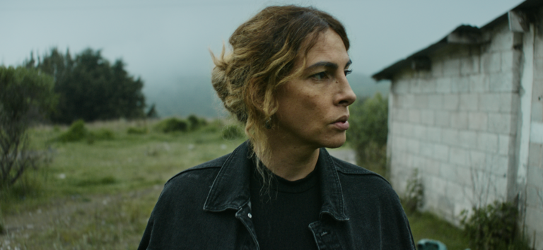 Woman with wavy brown hair in a black denim jacket looking sideways in an overcast rural landscape.