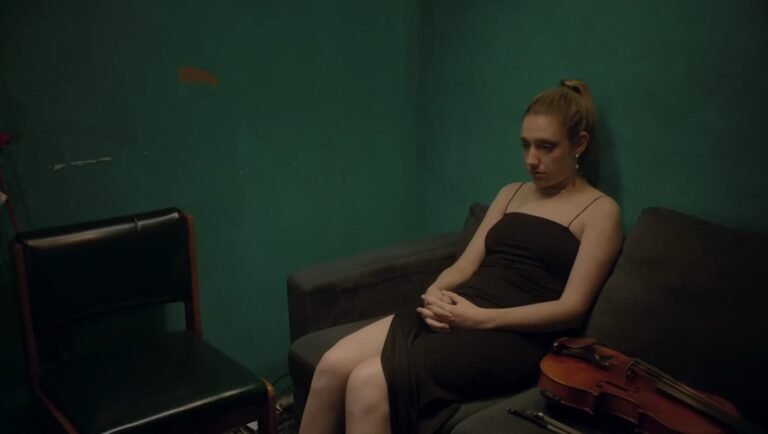 Isabel Hagen sits on a dark sofa against a green wall next to a violin with a bow.