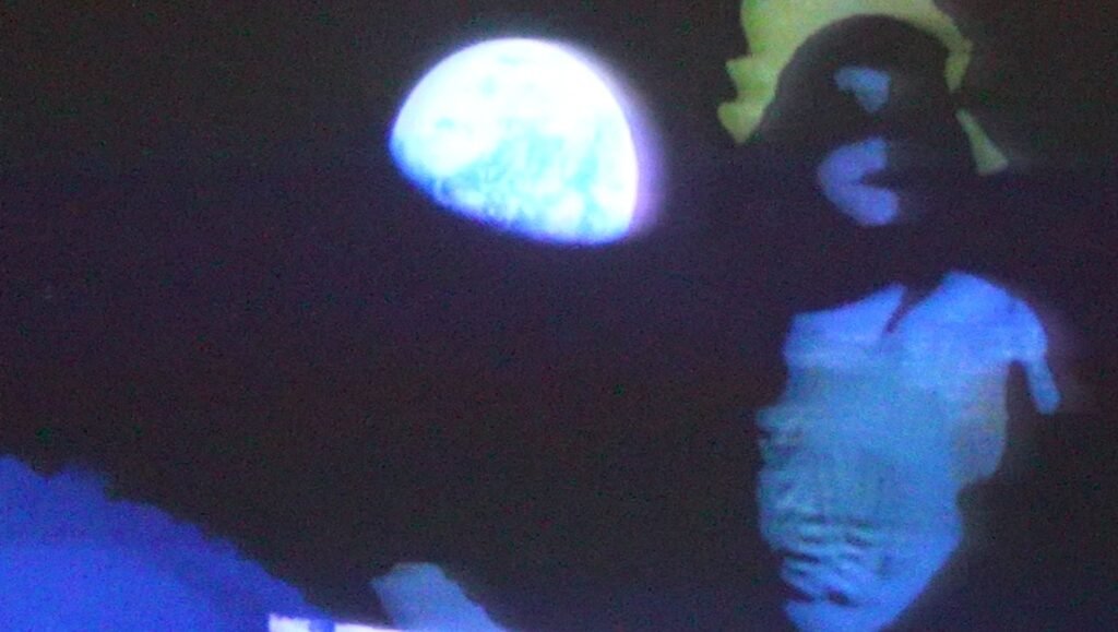 Glowing blue moon beside a shadowed figure in a yellow head covering against a dark night sky.