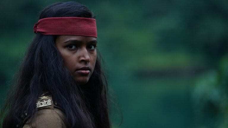 Woman with a red headband and long dark hair glancing sideways against blurred green foliage.