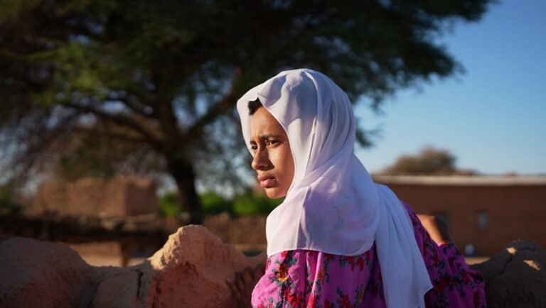 Woman in a white headscarf and floral pink dress gazing across a sunlit earthen wall with trees behind.