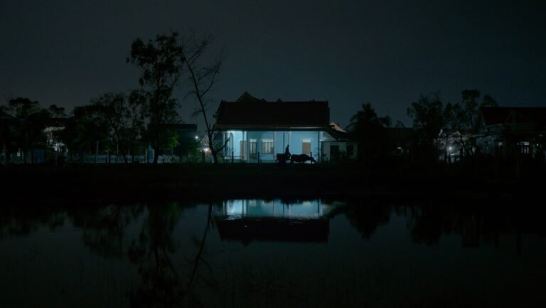 A stilt house with a pitched roof glows with blue light at night, reflected in dark water below trees.