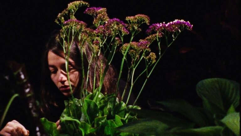 Woman with long dark hair looking down at tall green stems topped with clusters of small purple flowers against a black background