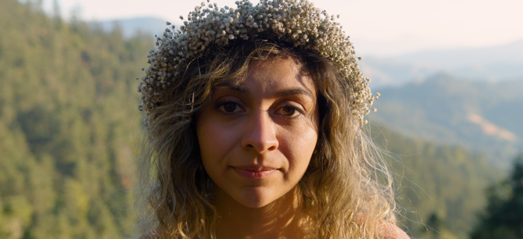 Woman with wavy hair and a dried flower crown looks ahead against a backdrop of green hills