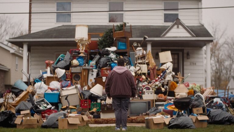 Person stands before a massive, overflowing pile of discarded furniture, household items, and trash in a front yard.