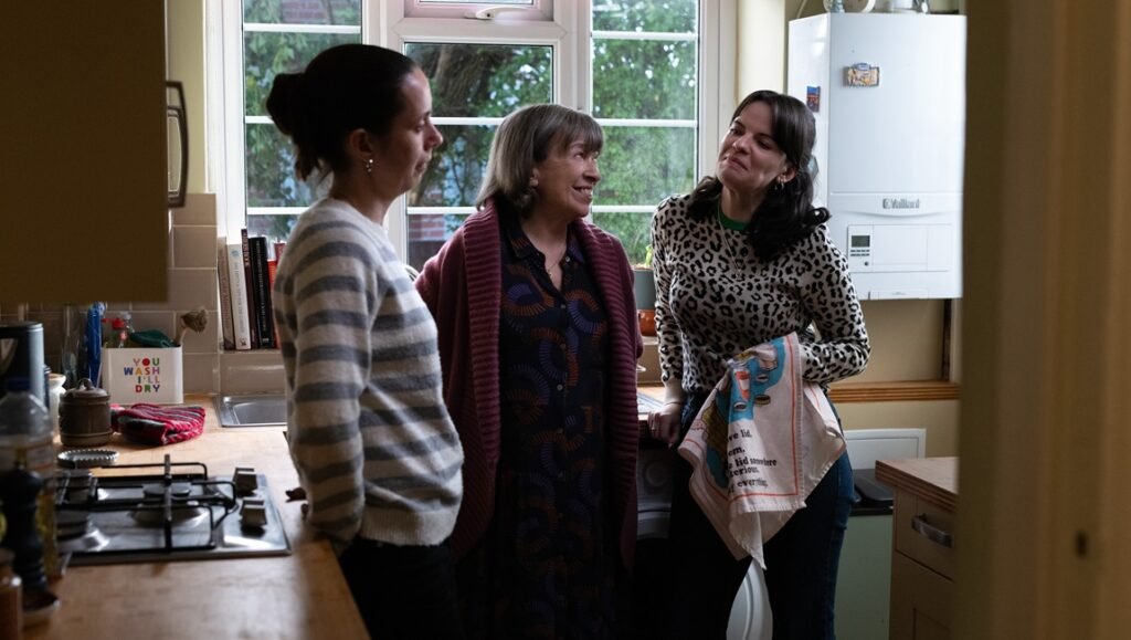 Three women talking in a kitchen, one holding a patterned tea towel