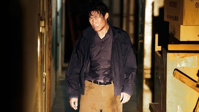 Man with a bruised face and disheveled hair standing in a dimly lit, narrow corridor wearing a dark jacket and tan pants.
