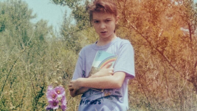 Young person in a lavender t-shirt clutching a rainbow-covered notebook while standing in a brushy, sunlit outdoor field.