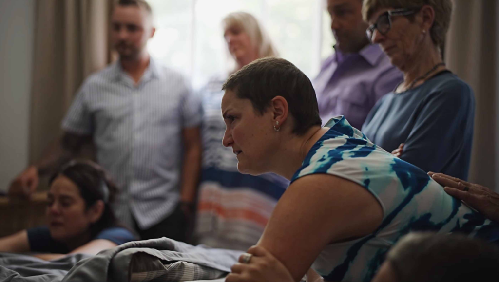 Short-haired woman in a blue and white patterned top leans forward intently as onlookers stand behind her.