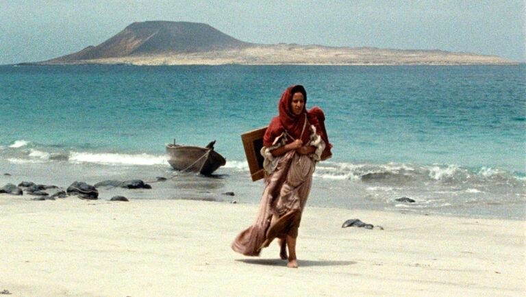 Woman in a red headscarf carries a framed picture and a child across a sandy beach toward turquoise water.