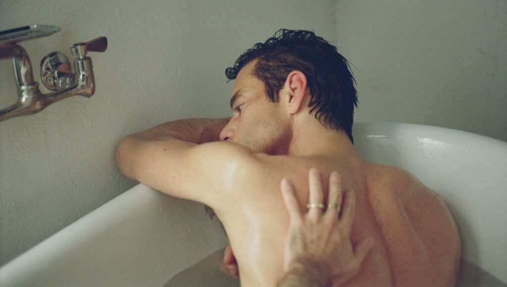 Shirtless man with wet hair leaning over a bathtub edge as a hand with a gold ring rests on his back.